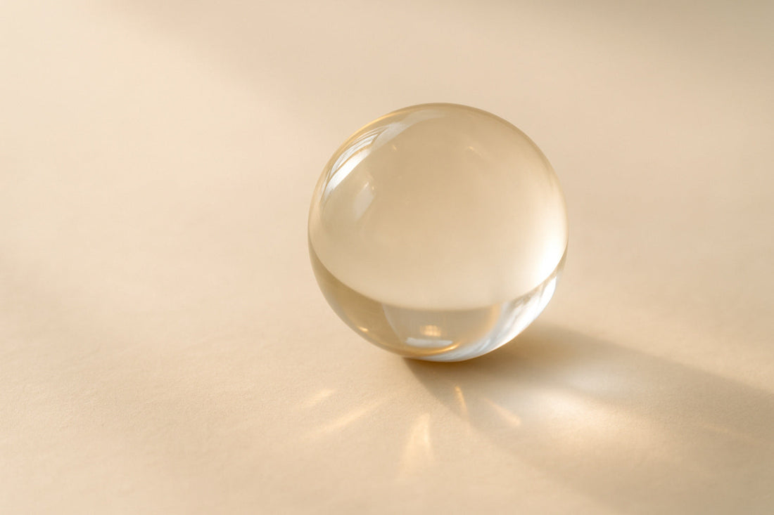 Eine transparente Glaskugel liegt im sanften, gold-weißen Licht auf heller Fläche. Licht und Schatten brechen sich auf ihrer Oberfläche – ein stilles Sinnbild für Zeit, Wahrnehmung und Balance.
