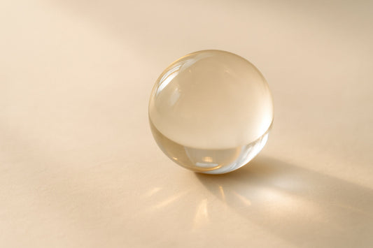 Eine transparente Glaskugel liegt im sanften, gold-weißen Licht auf heller Fläche. Licht und Schatten brechen sich auf ihrer Oberfläche – ein stilles Sinnbild für Zeit, Wahrnehmung und Balance.