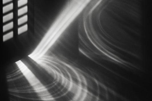 Schwarz-weißes Lichtmuster: Ein schmaler, präziser Lichtstrahl fällt durch Lamellen auf eine glatte Fläche und erzeugt klare, konzentrische Wellen aus Licht und Schatten.
