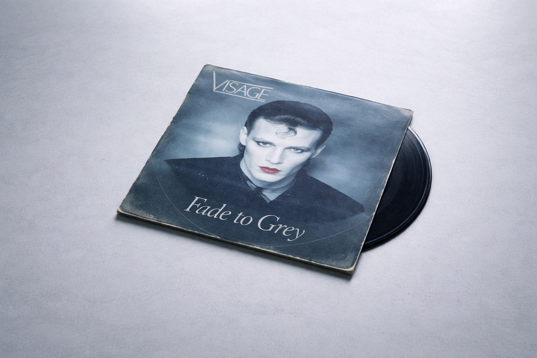 Eine leicht abgenutzte Vinyl-Single liegt ruhig auf einer hellgrauen Fläche. Das Cover von „Fade to Grey“ von Visage ist frontal leicht versetzt sichtbar, mit kühlen, entsättigten Farben. Gleichmäßiges Licht von oben