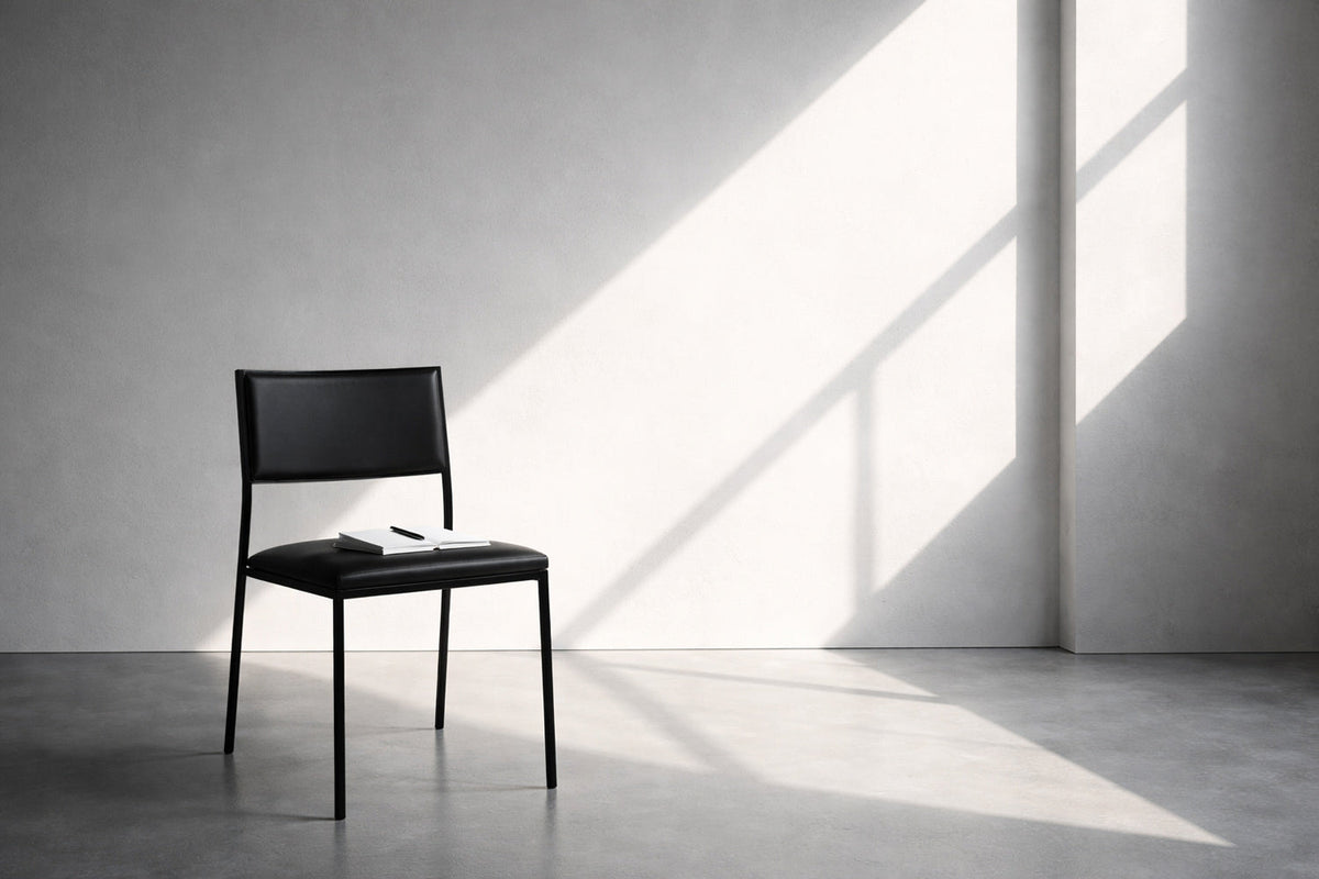 Karger, moderner Raum mit glatter Wand und scharfem Seitenlicht; ein schwarzer Stuhl mit Notizbuch steht allein im Raum, klare Schatten, ruhige Arbeitsatmosphäre.