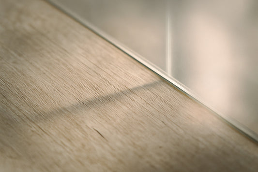Detailaufnahme einer Holzfläche mit einer Glaswand im sanften Seitenlicht – feine Maserungen, Lichtreflexe und weiche Schatten erzeugen ein stilles, poetisches Spiel aus Material, Zeit und Resonanz.