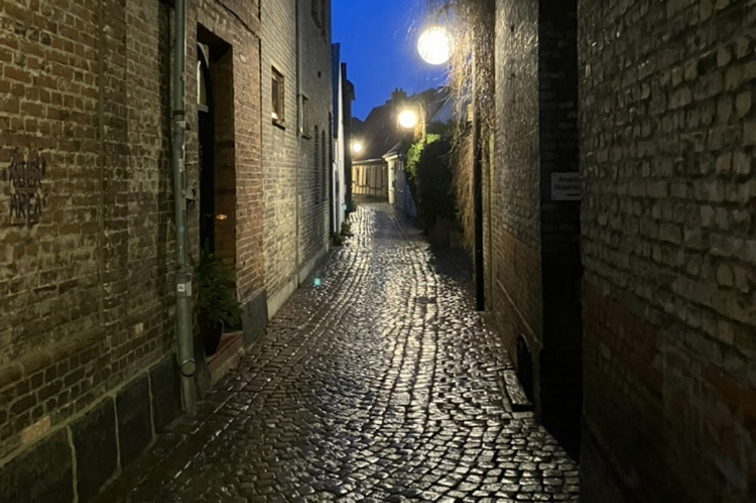 Schmale alte Gasse mit glänzendem Kopfsteinpflaster und warmen Laternen, Abendhimmel im Hintergrund. Ruhige, zeitvolle Stimmung zwischen Licht und Geschichte
