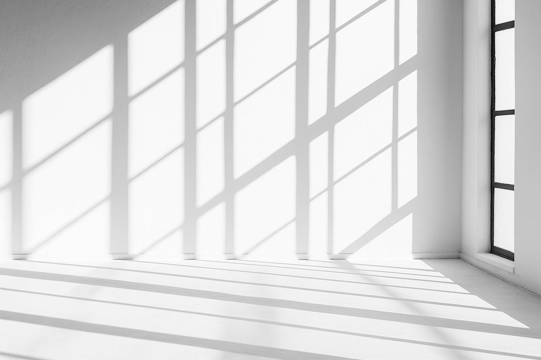 Sonnenlicht fällt durch hohe Fenster und wirft klare, geometrische Schatten auf eine weiße Wand und den Boden – ruhige, minimalistische Lichtstimmung.