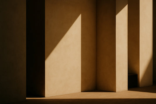Abstrakte architektonische Komposition in warmen, erdigen Tönen: mehrere sandfarbene Pfeiler werfen tiefe Schatten, während ein scharfes, diagonales Lichtdreieck über eine Wand gleitet.
