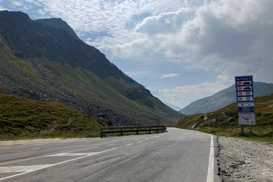 Bergpass mit sanfter Straße und Italia-Schild, weite Landschaft unter hellem Himmel – ein ruhiger Übergang zwischen zwei Ländern