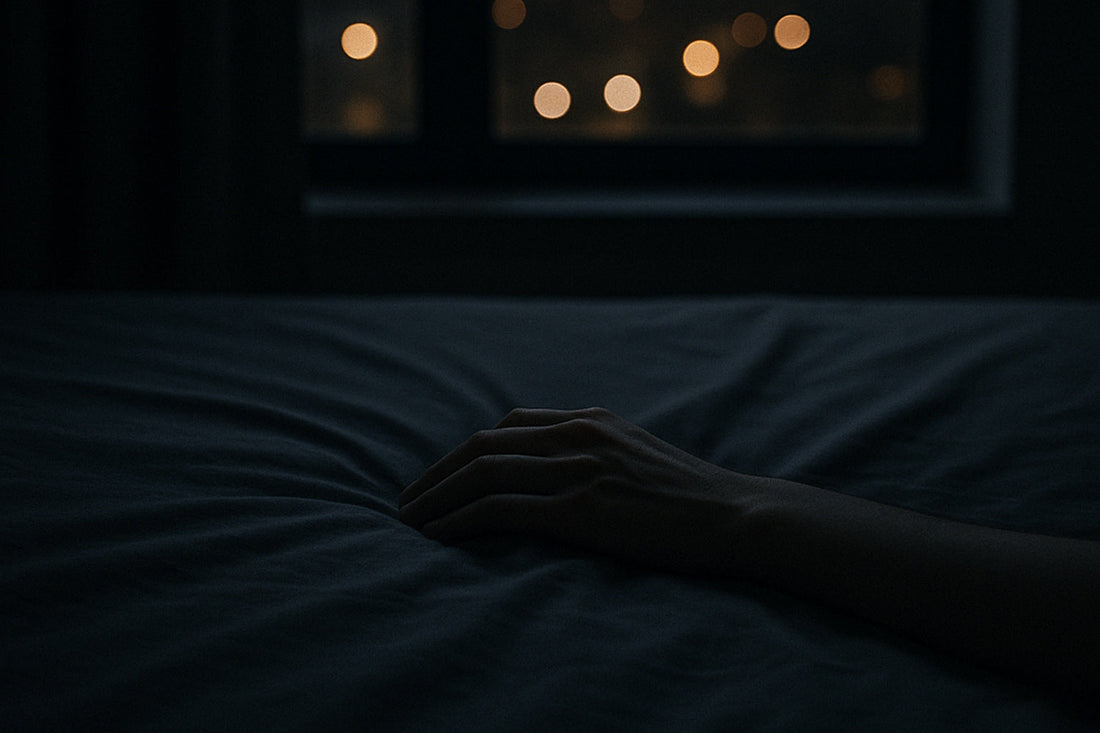 Eine Hand liegt auf einem Bettlaken, im Hintergrund unscharfe Stadtlichter durch ein Fenster – stille, urbane Szene über Träume und das innere Erleben der Nacht